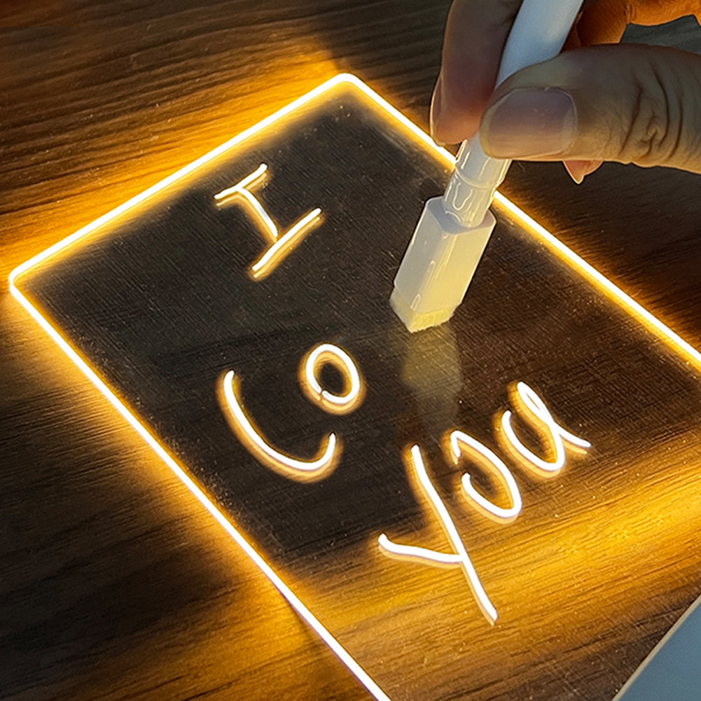 Glowing acrylic LED message board with handwritten notes using a white marker, used as a decorative night light on a wooden base.