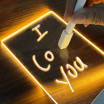 Glowing acrylic LED message board with handwritten notes using a white marker, used as a decorative night light on a wooden base.